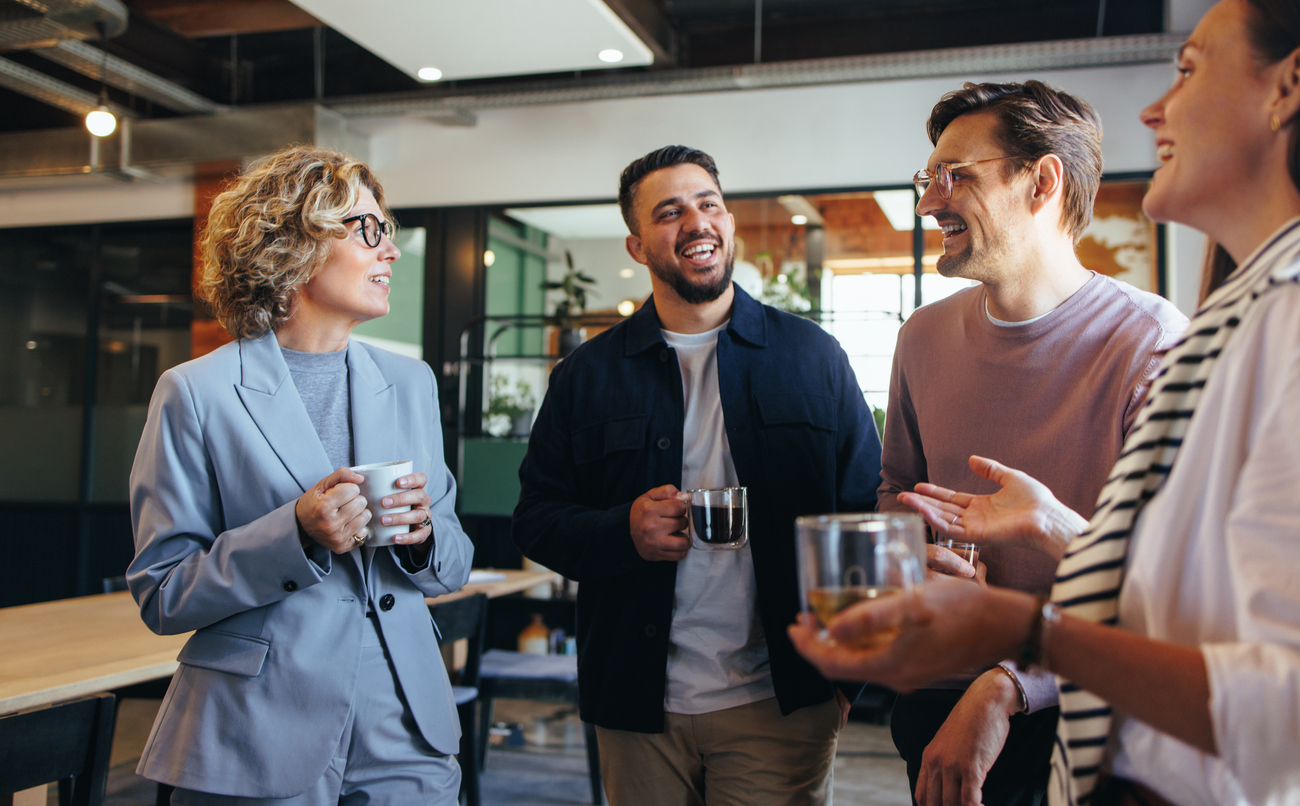 Vier collega’s praten en lachen tijdens een koffiepauze, als illustratie van sociaal contact en verbondenheid op het werk.