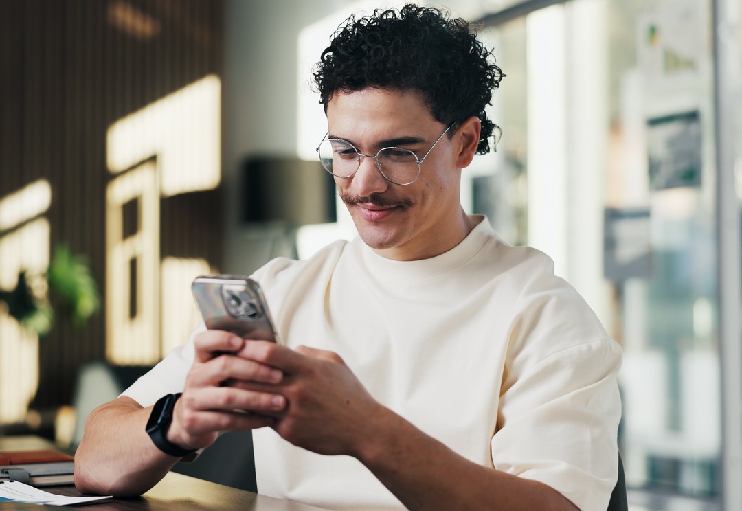 Man met een bril zit aan een bureau en kijkt naar zijn smartphone.